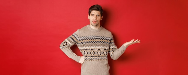 Image of disappointed and confused handsome man wearing christmas sweater raising hand and frowning ...