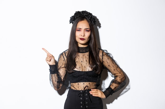 Image of disappointed and skeptical asian woman in witch costume complaining on something, pointing upper left corner and grimacing dissatisfied, standing over white background in halloween dress.