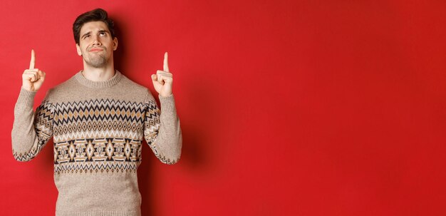 실망하고 회의적인 잘생긴 남자, 크리스마스 스웨터를 입고, 웃으면서 나쁜 것을 보고, 손가락을 가리키며, 빨간색 배경 위에 서 있는 것처럼 인상을 찌푸리는 이미지
