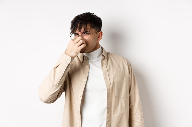 Image of disgusted guy shut his nose from awful smell, looking at something disgusting and stinky, standing on white background