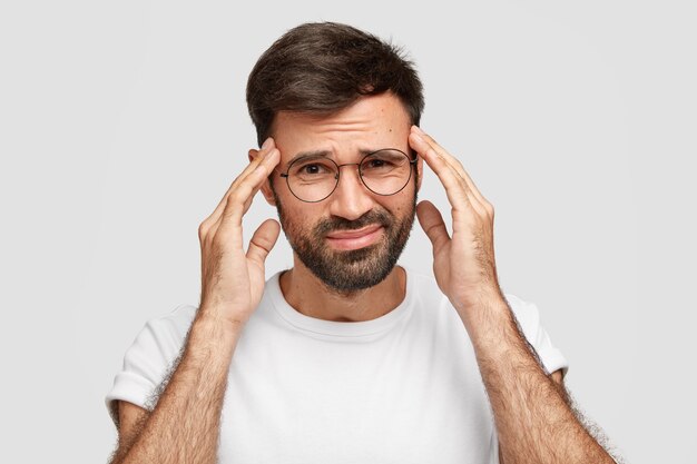 Image of displeased bearded male suffers from strong headache after working all night, has fatigue expression, keeps hands on temples, frowns face, poses against white wall. Bad feeling