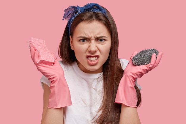 Image of displeased brunette housewife angry with much work about house, wears pink rubber gloves, clenches teeth in dislike