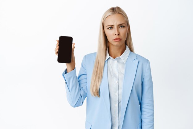 不機嫌そうな企業の女性が眉をひそめ、白い背景の上に青いスーツで立っている携帯電話の画面を表示している画像