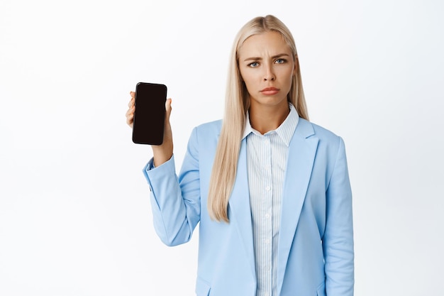 Изображение недовольной корпоративной женщины, хмурящейся и расстроенной, показывающей экран мобильного телефона, стоящего в синем костюме на белом фоне