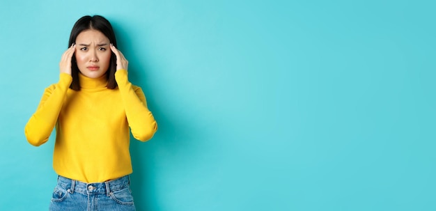 Free photo image of distressed asian woman touching head and frowning feeling headache standing troubled agains