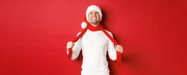 Image of distressed and frustrated man in santa hat chocking himself with scarf from sadness standin...