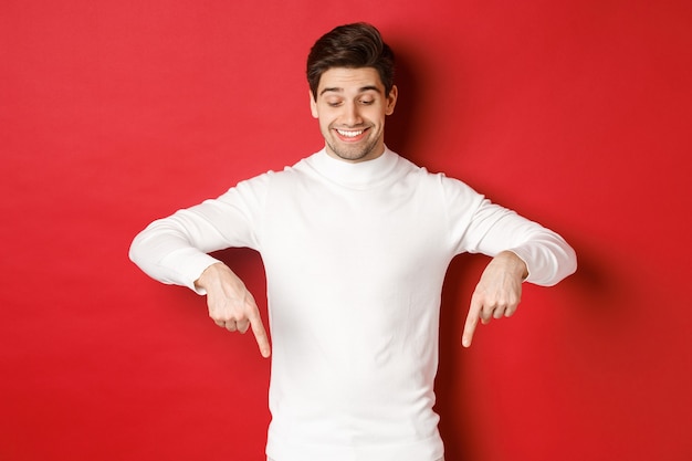 Изображение мечтательного красивого человека в белом свитере, смотрящего и указывающего пальцами на копировальное пространство, рекламы о новом году и праздниках, стоящего на красном фоне.