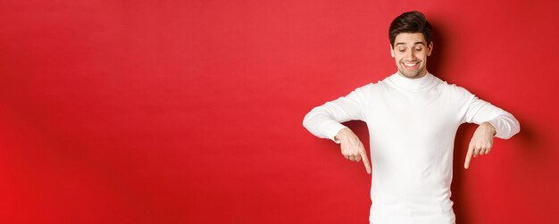 Image of dreamy goodlooking man in white sweater looking and pointing fingers down at copy space advertisement about new year and holidays standing over red background