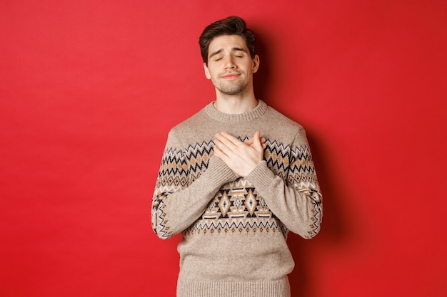 Free photo image of dreamy and happy, attractive bearded man in christmas sweater, holding hands on heart, smiling with eyes closed, remember something touching, celebrating new year, red background.