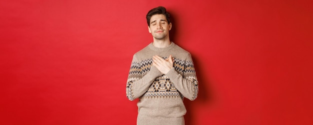 Image of dreamy and happy attractive bearded man in christmas sweater holding hands on heart smiling