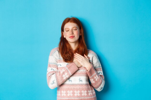 Image of dreamy redhead girl close eyes and holding hands on heart, feeling nostalgic, remember or imagine something, standing over blue background