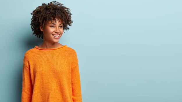 Image of dreamy woman with crisp hair looks pensively aside, has gentle smile on face, wears orange jumper, imagines something pleasant, models over blue wall, blank space for promotion