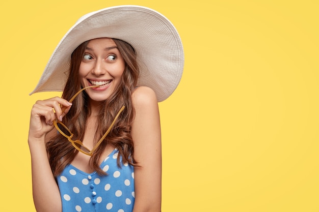  image of dreamy woman with long wavy hair, looks with pleased expression, has intention to travel abroad, holds sunglasses in hand, wears summer hat, free space for slogan over yellow wall