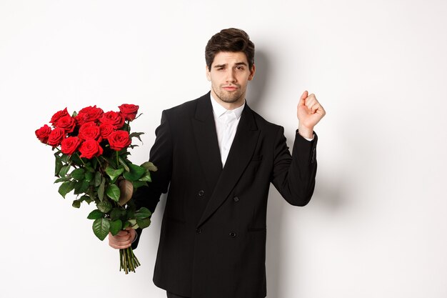 Image of elegant and sassy man in black suit, looking confident and holding bouquet of red roses, going on a romantic date, standing against white background