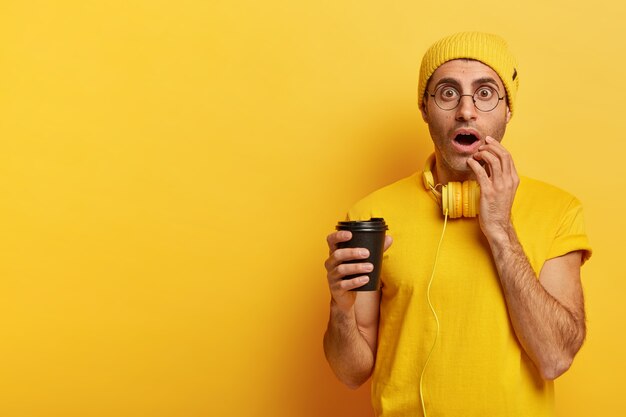 감성적 인 젊은이의 이미지가 일회용 커피 컵을 들고 눈을 크게 뜨고 눈을 믿을 수 없으며 노란 모자를 쓰고 있습니다.
