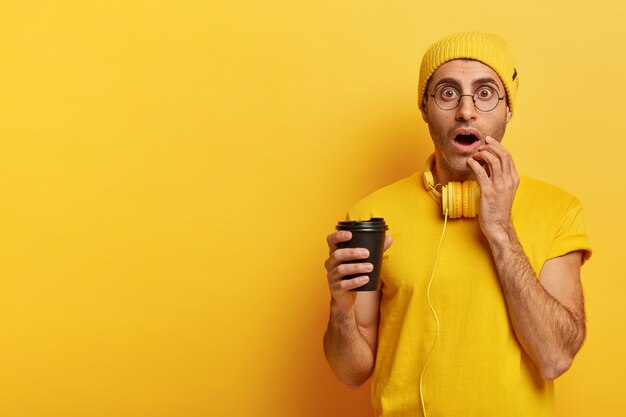 Image of emotional youngster holds disposable cup of coffee, stares with widely opened eyes, cannot believe eyes, wears yellow hat