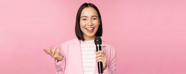 Image of enthusiastic asian businesswoman giving speech talking with microphone holding mic standing in suit against pink studio background