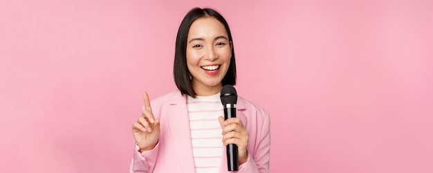 Изображение восторженной азиатской бизнесвумен, выступающей с речью, разговаривающей с микрофоном, держащей микрофон, стоящей в костюме на фоне розовой студии