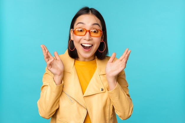 Image of enthusiastic young asian woman celebrating triumphing looking surprised and happy clapping hands satisfied standing over blue background