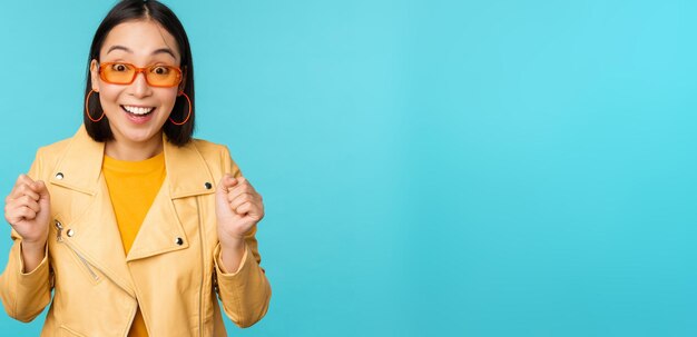 Image of enthusiastic young asian woman celebrating triumphing looking surprised and happy clapping hands satisfied standing over blue background