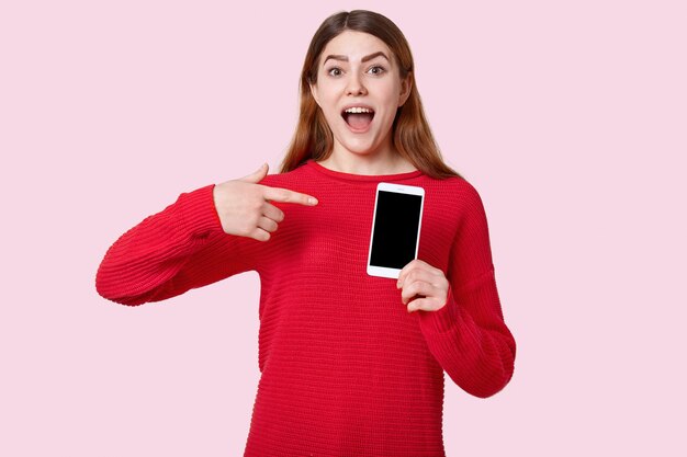 Изображение европейской позитивной молодой женщины указывает на сотовый телефон с пустым экраном, одетый в красный свитер, рекламирует новый гаджет