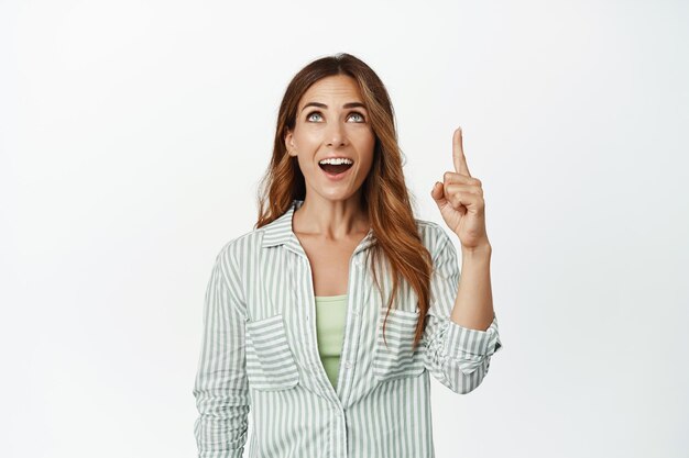 Image of excited, amazed brunette woman, gasp with WOW face, pointing finger and looking up at smth awesome, like promo deal, standing against white background