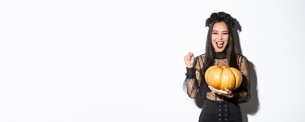 Free photo image of excited beautiful asian woman celebrating halloween wearing witch outfit and holding pumpki