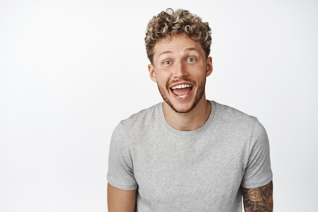 Image of excited blond guy smiling looking amazed and happy at camera hear big announcement standing in gray tshirt over white background