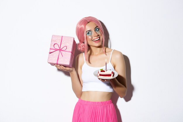 Изображение возбужденной милой девушки в розовом парике, трясущейся коробкой с подарком и блуждающей внутри, держащей в руках кусок торта на день рождения, празднующей день рождения.