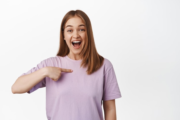 Image of excited girl points at herself and smiles happy, being a winner, receive big good news, rejoicing was chosen, standing over white background