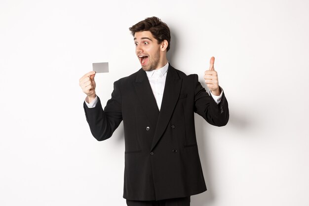 Image of excited handsome businessman, showing credit card and thumb-up, standing against white background