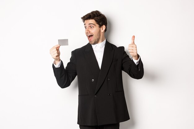 Изображение возбужденного красивого бизнесмена, показывающего кредитную карту и большой палец вверх, стоящего на белом фоне