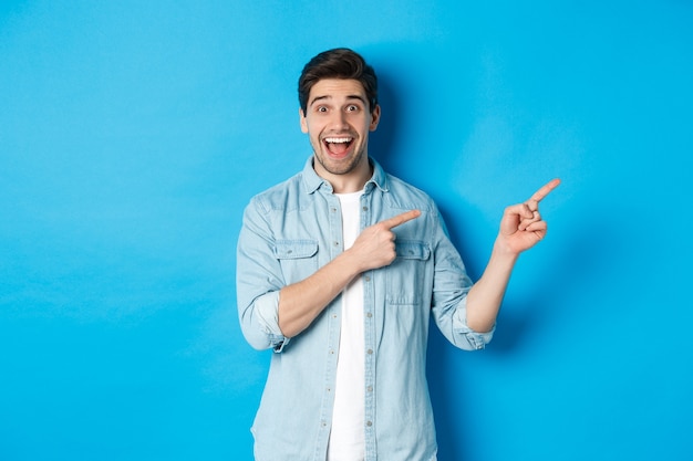 캐주얼 복장을 한 흥분한 잘생긴 남자의 이미지, 광고를 보여주고, 복사 공간을 손가락으로 가리키며 웃고, 파란색 배경에 서 있는 이미지