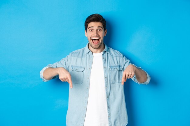 Image of excited handsome man pointing fingers down, making an announcement, standing against blue background