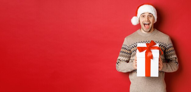 Immagine di un bell'uomo eccitato che riceve un regalo di natale, indossa un cappello da babbo natale e un maglione invernale, grida di gioia, tiene un regalo e sta in piedi su sfondo rosso