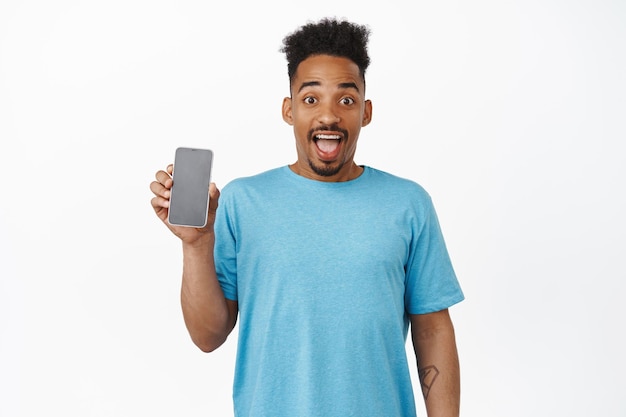 Image of excited happy guy, african american boy showing smartphone screen, app on mobile phone, scream from amazement, standing in blue t-shirt over white background.