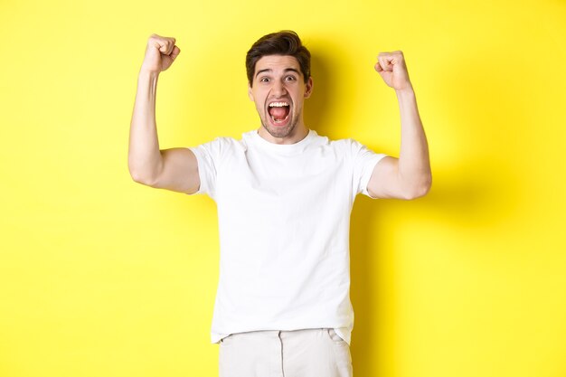 Image of excited man winning, raising hands up and celebrating, triumphing and rooting for team, standing over yellow background. Copy space