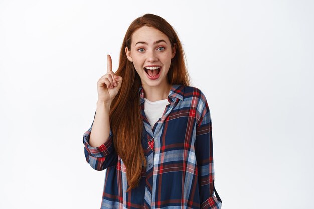Image of excited redhead girl got an idea, here is solution, raising finger with amazed face, got plan, stands against white background