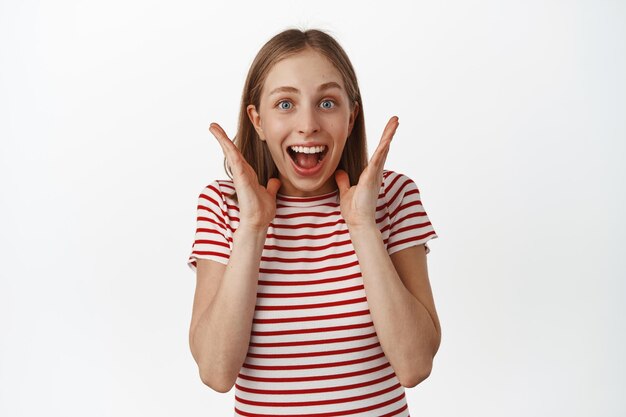 Image of excited and surprised young woman rejoice over big surprise, hold hands near face, shouting and smiling amazed, celebrating, receive great news, standing against white background.