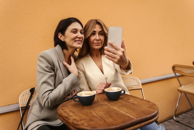 コーヒーを飲みながら自撮り写真を撮るカフェで屋外に座っている色白の大人のガールフレンドの画像携帯電話のコンセプト