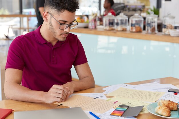 Image of focused male student prepares report in finances, looks attentively at papers, eats delicious croissants, poses over cafe interior with free space for your promotion. Freelance work