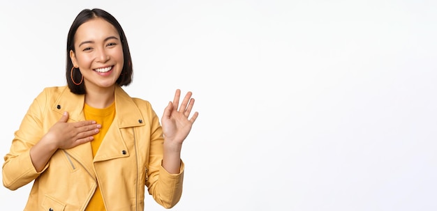 Image of friendly asian girl in stylish yellow coat raising arm introduce herself greeting waving hand saying hello standing over white background