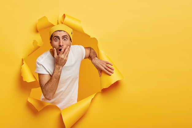 Image of frightened puzzled guy covers mouth, stares with scared expression, cannot believe his eyes