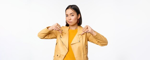 Image of fully disappointed asian woman showing thumbs down shaking head displeased dislike smth standing over white background