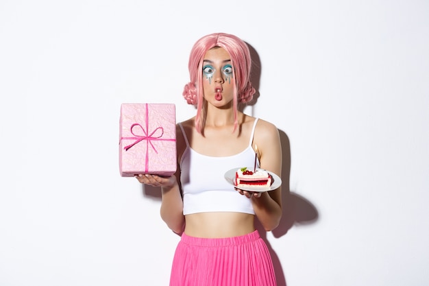 Изображение смешной именинницы, корчащей глупые рожи, держа в руках торт и упакованный подарок, стоя.