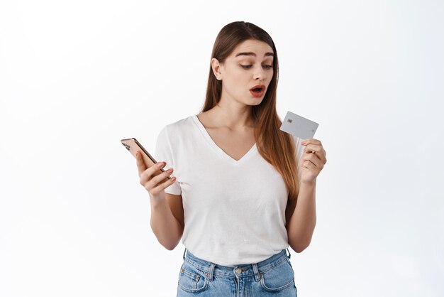 Image of girl looks curious at bank credit card holds smartphone in hand pays for online order shopping in internet store standing over white background