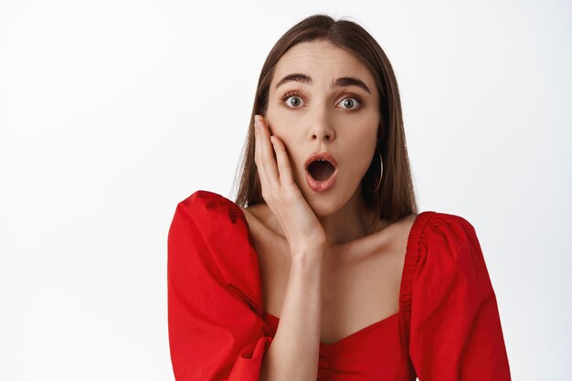 Image of girl in red dress, look amazed and surprised, open mouth and gasp thrilled, hear amazing news, big promo deal, standing in disbelief against white background.