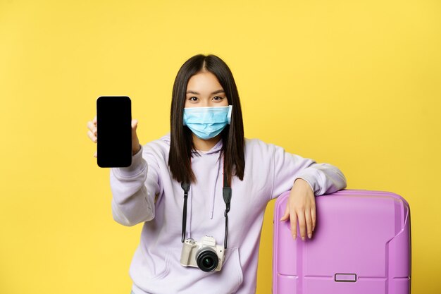 Изображение девушки-путешественницы, азиатской туристки показывает экран своего телефона, паспорт здоровья covid в приложении для смартфона, в медицинской маске и чемодане, желтый фон.