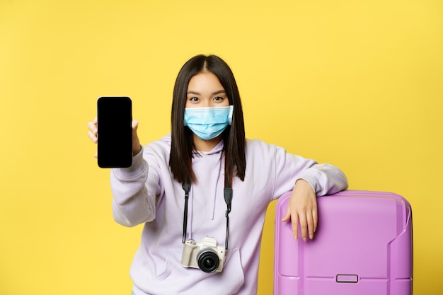 女の子の旅行者、アジアの観光客の画像は、彼女の電話スクリーン、スマートフォンアプリでの健康パスポート、医療用マスクとスーツケース、黄色の背景を示しています。