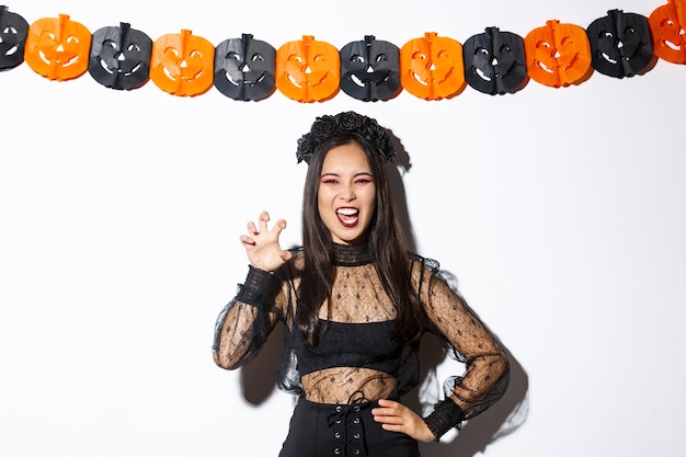 Immagine della ragazza in costume da strega cattiva che agisce male e spaventosa, facendo smorfie su sfondo bianco con decorazioni di halloween.